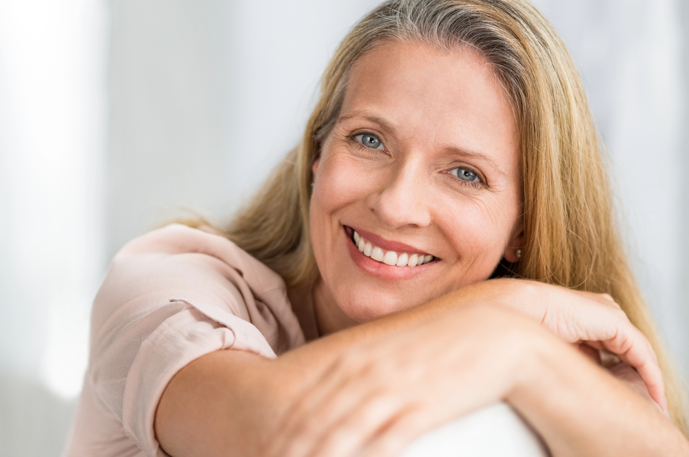 A woman with long blonde hair and veneers smiles while resting her arms on a white surface; she is wearing a light-colored top.