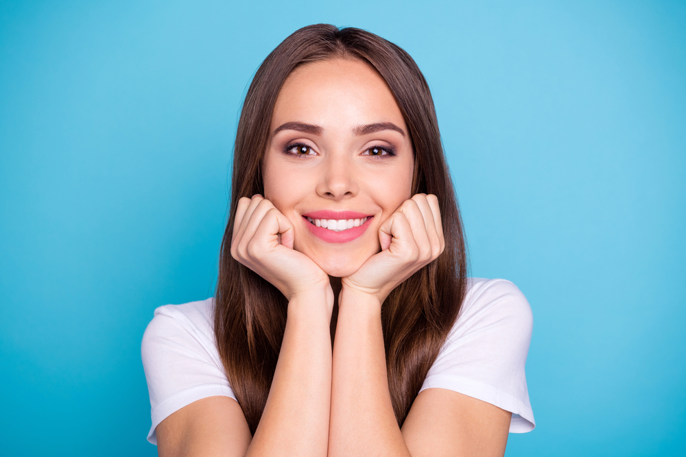 A woman with long brown hair smiles, resting her chin on her hands. She is wearing a white shirt and is positioned against a blue background.