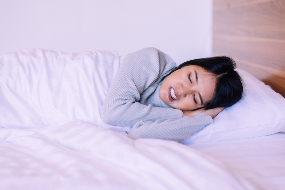 A person with short black hair, wearing a gray long-sleeve shirt, lies on their side in bed with white sheets. They appear to be asleep, resting their head on a pillow and smiling slightly.