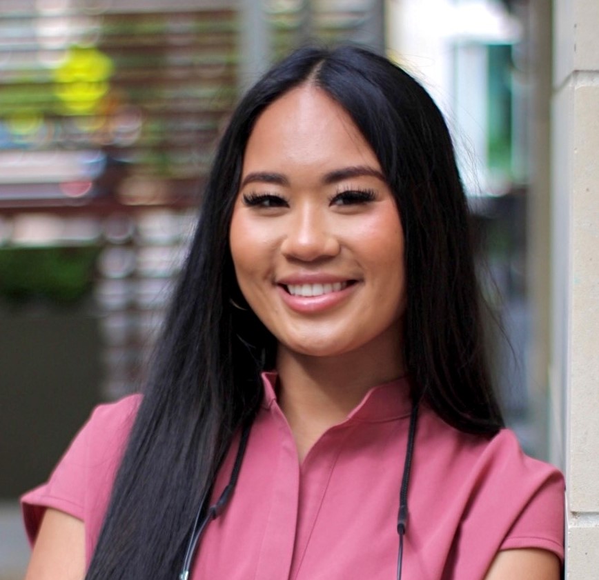 A person with long black hair, wearing a pink top, smiles while standing outdoors. The background is blurred, showing some greenery and buildings.