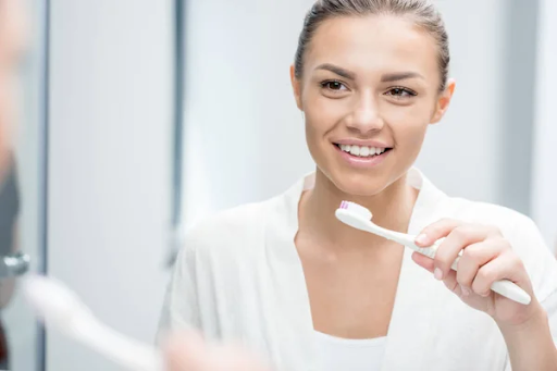 A person smiling in a bathroom holds a toothbrush with toothpaste, wearing a white robe and looking into the mirror. The background is softly blurred, enhancing the serene setting.